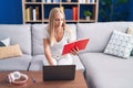 Young blonde woman student sitting on sofa reading book at home Royalty Free Stock Photo