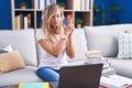 Young blonde woman studying using computer laptop at home holding symbolic gun with hand gesture, playing killing shooting