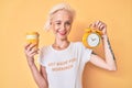 Young blonde woman with tattoo holding alarm clock and take away cup of coffee winking looking at the camera with sexy expression, Royalty Free Stock Photo