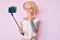 Young blonde woman with tattoo taking a selfie photo with smartphone covering mouth with hand, shocked and afraid for mistake