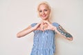 Young blonde woman with tattoo wearing casual clothes smiling in love doing heart symbol shape with hands