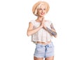 Young blonde woman with tattoo wearing summer hat begging and praying with hands together with hope expression on face very