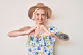Young blonde woman with tattoo wearing summer hat smiling in love showing heart symbol and shape with hands