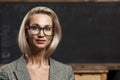 A young blonde woman teacher with glasses stands in a class near a black school board. Distance learning. Close-up. Space for text Royalty Free Stock Photo