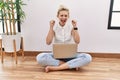 Young blonde woman using computer laptop sitting on the floor at the living room excited for success with arms raised and eyes Royalty Free Stock Photo