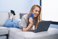 Young blonde woman using laptop and credit card lying on sofa at home Royalty Free Stock Photo