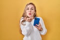 Young blonde woman using smartphone typing message looking at the camera blowing a kiss with hand on air being lovely and sexy Royalty Free Stock Photo