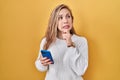 Young blonde woman using smartphone typing message thinking worried about a question, concerned and nervous with hand on chin Royalty Free Stock Photo