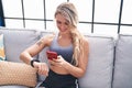 Young blonde woman using smartphone and watch sitting on sofa at home Royalty Free Stock Photo