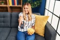 Young blonde woman watching tv eating chips potatoes sitting on sofa at home Royalty Free Stock Photo