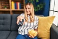 Young blonde woman watching tv eating chips potatoes sitting on sofa at home Royalty Free Stock Photo