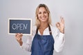 Young blonde woman wearing apron holding blackboard with open word doing ok sign with fingers, smiling friendly gesturing Royalty Free Stock Photo