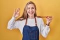 Young blonde woman wearing apron tasting food holding wooden spoon winking looking at the camera with sexy expression, cheerful Royalty Free Stock Photo