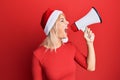 Young blonde woman wearing christmas hat screaming using megaphone over isolated red background Royalty Free Stock Photo