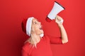 Young blonde woman wearing christmas hat screaming using megaphone over isolated red background Royalty Free Stock Photo