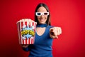 Young blonde woman wearing 3d glasses and eating pack of popcorn watching a movie on cinema pointing to you and the camera with Royalty Free Stock Photo