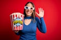 Young blonde woman wearing 3d glasses and eating pack of popcorn watching a movie on cinema Waiving saying hello happy and Royalty Free Stock Photo