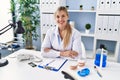 Young blonde woman wearing dentist uniform sitting on table at clinic