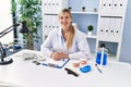 Young blonde woman wearing dentist uniform sitting on table at clinic