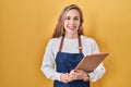 Young blonde woman wearing professional waitress apron holding clipboard looking positive and happy standing and smiling with a