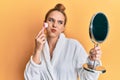 Young blonde woman wearing robe holding makeup remover brush puffing cheeks with funny face