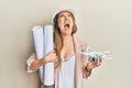 Young blonde woman wearing safety helmet holding blueprints and drone angry and mad screaming frustrated and furious, shouting Royalty Free Stock Photo