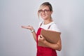 Young blonde woman wearing waiter uniform holding clipboard pointing aside with hands open palms showing copy space, presenting Royalty Free Stock Photo