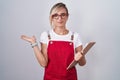 Young blonde woman wearing waiter uniform holding clipboard smiling cheerful presenting and pointing with palm of hand looking at Royalty Free Stock Photo