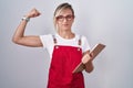 Young blonde woman wearing waiter uniform holding clipboard strong person showing arm muscle, confident and proud of power