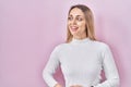 Young blonde woman wearing white sweater over pink background looking away to side with smile on face, natural expression Royalty Free Stock Photo