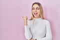 Young blonde woman wearing white sweater over pink background smiling with happy face looking and pointing to the side with thumb Royalty Free Stock Photo