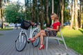 A young blonde woman with a white bicycle is sitting on a bench and reading a book - image Royalty Free Stock Photo