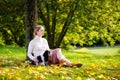 A young blonde woman in a white sweater is sitting near a tree in an autumn sunny park with her Cavalier King Charles Spaniel dog