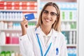 Young blonde woman working at pharmacy drugstore holding credit card looking positive and happy standing and smiling with a Royalty Free Stock Photo