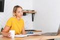 Young blonde woman in yellow t-shirt working at laptop in kitchen at home, girl with cup of the morning coffee with papers Royalty Free Stock Photo