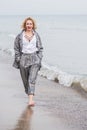 A young blonde of 30 years old walks on the white beach sand and keeps her hands in her pants pockets, the sea wave raises foam.