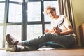 Young blondhair handsom man plays on guitar sitting on floor near the whole wall window at home. Music education concept image. Royalty Free Stock Photo