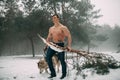 Young bodybuilder with bare torso leads dog Malamute and carries pine branch in his hand at walk in winter forest.