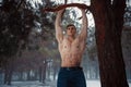 Young bodybuilder with bare torso pulls up on branch in forest in winter. Body hardening.