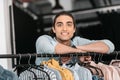 Young boutique owner leaning at hangers with clothes and smiling at camera