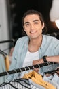 Young boutique owner leaning at hangers with clothes and smiling at camera