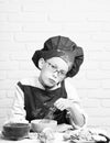 Young boy cute cook chef in red uniform and hat on stained face with glasses sitting on table with colorful bowls, tasty
