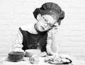 Young boy cute cook chef in red uniform and hat on stained face with glasses sitting on table with colorful bowls, tasty