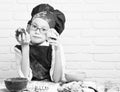 Young boy cute cook chef in red uniform and hat on stained face with glasses sitting on table with colorful bowls, tasty