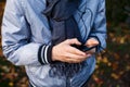 Young boy in gray jacket with gray scarf holds and uses smartphone with headphones outside over autumn background. Technology