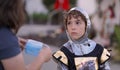 A young boy in a Halloween costume looks unhappy and questioning as his mother puts on a face mask for safety and protection.