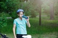 Young boy in helmet and green t shirt cyclist drinks water from bottle in the park. Smiling cute Boy on bicycle in the forest