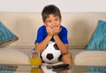 Young boy holding soccer ball watching excited and nervous football game on television biting fingernails sitting at living sofa c