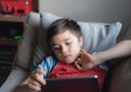 Young boy holding tablet looking out deep in thought, Unhappy kid with bored face sitting alone on sofa, A lonely child playing Royalty Free Stock Photo