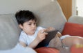 Young boy holding tablet looking out deep in thought, Unhappy kid with sad face sitting alone on sofa, A lonely child playing with Royalty Free Stock Photo